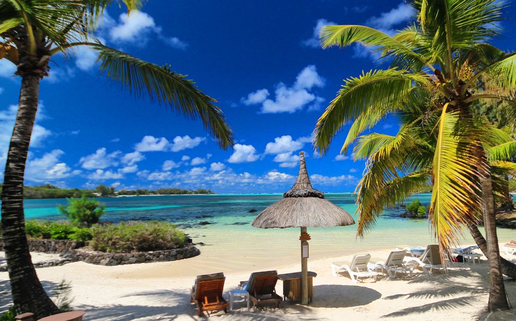 Playa de arena blanca en isla Mauricio con agua turquesas, palmeras, una sombrilla y varias tumbonas. 